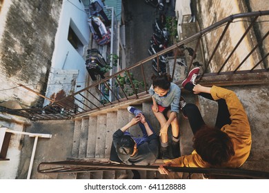 High Angle View Of Stylish Asian Teenagers Hanging Out In Urban Slums: They Sitting On Stairs Of Shabby Apartment House And Talking To Each Other