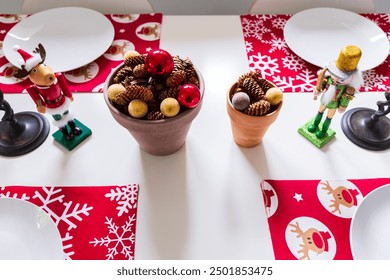 High angle view of table being set for Christmas lunch with nutcracker soldier and moose figurines, cute red table mats and terracotta pots filled with acorns and shiny balls - Powered by Shutterstock