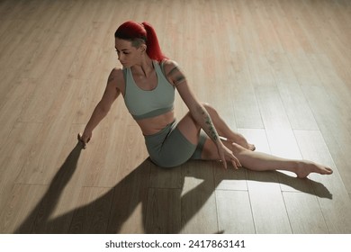 High angle view of tattooed Caucasian girl with red ponytail stretching hips sitting on studio floor in dramatic lighting - Powered by Shutterstock