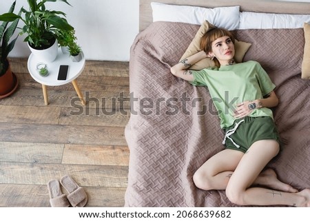 high angle view of tattooed woman lying on bed near bedside table with plants and mobile phone