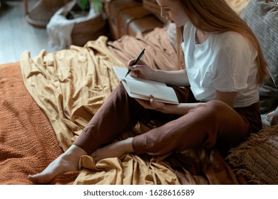 High Angle View Of Thoughtful Young Adult Girl Writing Notes In Diary, Sitting On Bed, Resting In Home At Cozy Bedroom In Boho Style