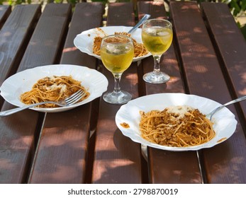 High angle view of three plates of delicious spaghetti bolognese with grated cheese and two glasses of chilled white wine served on a wooden picnic table under dappled sunlight