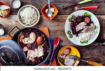 High Angle View of Traditional Brazilian Stew Made with Beans and Meats Served on Rustic Wooden Table with Side Plates, Rice and Garnishes
