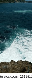 High angle view of turquoise ocean waves crashing against a rocky cliff, creating white sea foam