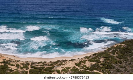 High angle view of turquoise waves crashing onto a sandy beach with coastal vegetation.