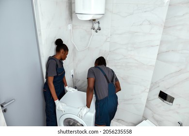 High Angle View Two African Man And Woman In Work Clothes Moving Washing Machine In White Bathroom. Moving Company Staff. Black And White Colours Contrast.