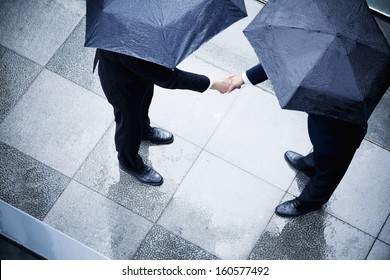 High angle view of two businessmen holding umbrellas and shaking hands in rain - Powered by Shutterstock