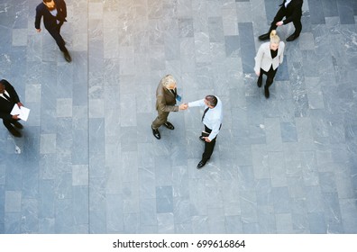 High Angle View Of Two Businessmen Shaking Hands Together While Colleagues Walk Around Them In The Lobby Of A Busy Modern Office Building