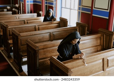 High angle view of two diverse Catholic nuns sitting on pews in church, praying with rosary beads in hands, copy space