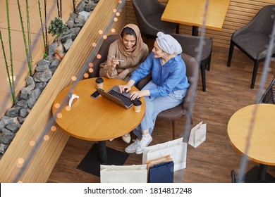 High angle view at two Middle-Eastern young women enjoying online shopping via laptop while sitting in mall cafe and holding credit card, copy space