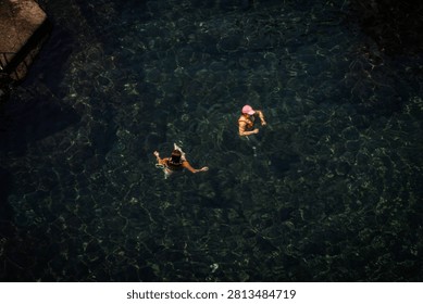 High Angle View of Two People Swimming in Clear Dark Ocean Water Near Coastal Rocks