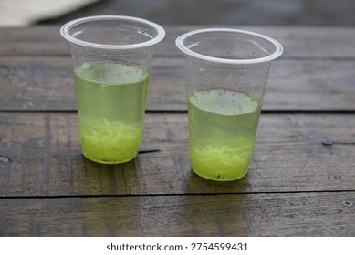 High angle view of two plastic cups containing fresh lime or cucumber drinks with shredded fruit inside on a weathered wooden table.
