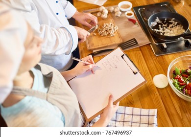 High angle view of unrecognizable woman wearing apron standing next to chef and taking necessary notes while he showing how to prepare salad with mushrooms - Powered by Shutterstock