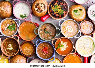High Angle View of Various Comforting and Savory Gourmet Soups Served in Bread Bowls and Handled Dishes and Topped with Variety of Garnishes on Table Surface with Gray Tablecloth