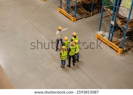 High angle view of a warehouse manager giving instructions and explaining logistics to a group of workers wearing safety vests and helmets during a briefing inside a large distribution center