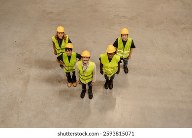 High angle view of warehouse workers team wearing safety vests and helmets standing together in large distribution warehouse, demonstrating teamwork and workplace safety