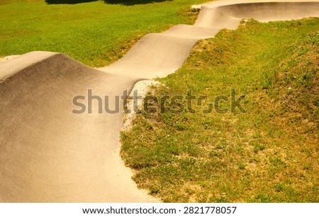 High angle view of a wavy asphalt section of a pump track circuit with smooth turns and bumps running through green grass in an outdoor sports park. Perfect for commercial graphics, sports facility marketing, urban infrastructure projects, cycling, skateboarding culture, and modern park design concepts.