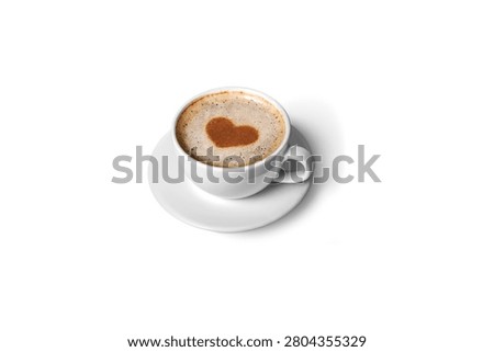 High angle view of a white ceramic cup and saucer filled with hot cappuccino, latte, or macchiato, decorated with a sweet brown heart shape made from cocoa powder or cinnamon stenciled onto the milk foam. Isolated on a solid white background.