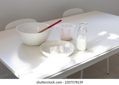 High Angle View Of White Cooking Utensils And Bowl, Milk, Eggs And Flour In Measuring Cup Set On Table In Dappled Light