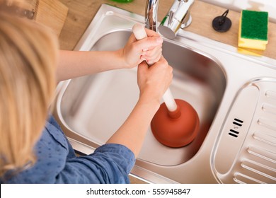 High Angle View Of Woman Using Plunger In Blocked Kitchen Sink To Unclog Drain