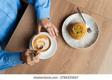High angle view of a woman's hands stirring a cappuccino next to a sweet pistachio pastry on a wooden table