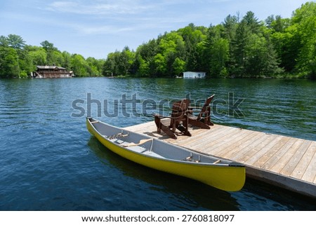 High angle view of a yellow canoe is moored alongside a weathered dock in Muskoka, Ontario, Canada, where two dark wood Adirondack chairs face the calm lake. Two paddleboarders can be seen near the cottages on the far shore, surrounded by lush summer foliage beneath a partly cloudy blue sky — a quintessential Canadian cottage country scene.