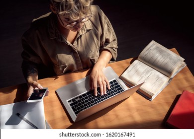 High Angle View Of Young Adult Woman Spending Free Time At Student Library, Preparation To Examination. Woman Sitting Behind Table, Using Laptop Computer And Modern Smartphone