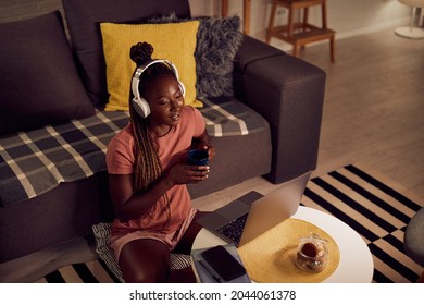 High angle view of young African American woman with headphones using laptop and drinking tea in the evening at home. - Powered by Shutterstock