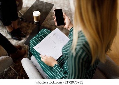 High Angle View Of Young Businesswoman With Blond Hair Holding Report And Using Smartphone While Sitting On Chair In Office