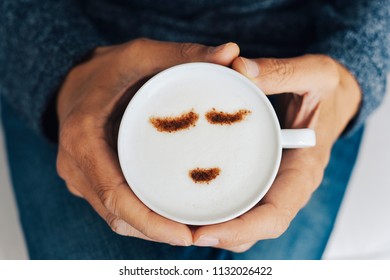 high angle view of a young caucasian man having a cup of cappuccino in his hands, with a happy face drawn with cocoa powder on its milk foam