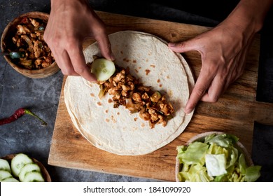 high angle view of a young caucasian man preparing a durum or a burrito, with chicken meat cooked with different vegetables such as onion or red and green pepper, and fresh lettuce and raw cucumber