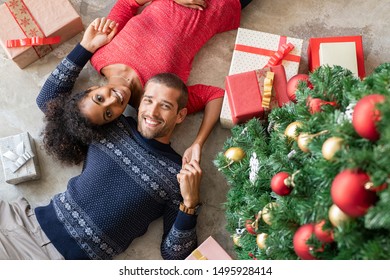 High Angle View Of Young Man And African Woman Lying On Floor Near Christmas Tree With Presents. Top View Of Happy Multiethnic Couple Lying Down Near Xmas Gifts Holding Hands And Looking At Camera.