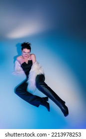 High Angle View Of Young Model In White Faux Fur Jacket And Black Leather Pants Posing On Blue Background