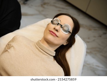 High angle view of a young pretty European woman wearing UV protective goggles and lying down on the daybed in a wellness spa clinic. Face Care. Facial Laser Hair Removal. Epilation. Smooth Skin.