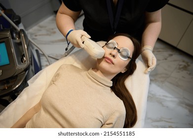 High angle view of a young pretty Caucasian middle-aged woman wearing UV protective goggles, receiving facial laser treatment and lying down on the daybed in a wellness spa clinic