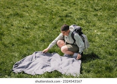 High angle view of young short haired female hiker with fitness tracker, backpack and travel equipment putting blanket on grassy lawn, connecting with nature concept, summer