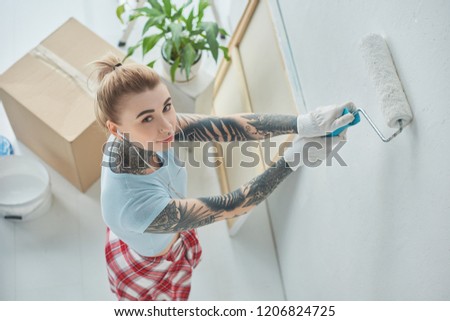 high angle view of young tattooed woman in earphones painting wall at new home