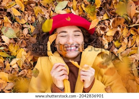 High angle view of young woman lying over yellow leaves. Beautiful woman laughing and throwing leaves while lying on ground. Top view of cheerful girl surrounded by autumn leaves at park.