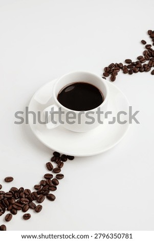 High angle of white ceramic cup filled with black coffee on saucer, with roasted coffee beans arranged in a diagonal curve on a clean white background.