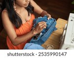 High angle of young female worker of thrift shop fixing pearl beads to secondhand denim jacket while sitting by workplace