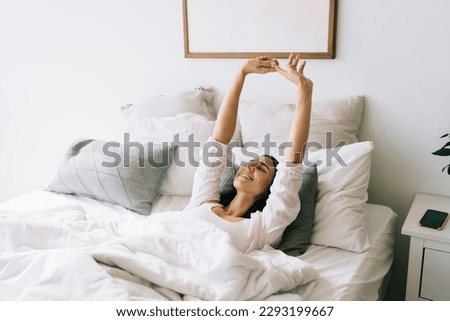High angle of young happy female in pajamas lying in bed with cushions smiling and stretching with eyes closed after awakening early in morning