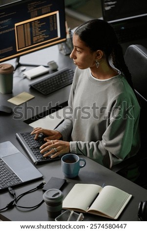 High angle of young Middle Eastern female programmer immersed in coding on keyboard of desktop PC, while working at cluttered with stationery and cups desk in office