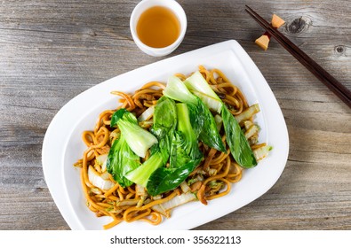 High Angled View Of Spicy Cooked Noodles, Onion, Bok Choy, Chicken Slices, Chopsticks And Tea On Rustic Wood. 