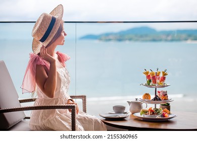 High Class Tea With Selection Of Various Desserts In Luxury Restaurant With Perfect Sea View. Elegant Woman In White Dress, Straw Hat Enjoy Sweet Treats From Decorated Stand With Pastry. Slow Motion