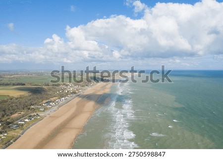 A high drone perspective captures the long sandy stretch of Omaha Beach bordered by gentle waves, green fields, and scattered houses under a sky filled with dramatic cumulus clouds.