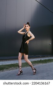 High End Fashion Image Of Tall Slim Brunette Model Wearing Black Leather Dress, High Heels Summer Shoes. Statement Sequins Earrings And Bright Vivid Brown Make Up, Bun Hairstyle. Outdoor Photo Shoot