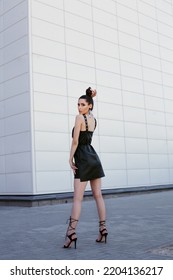 High End Fashion Image Of Tall Slim Brunette Model Wearing Black Leather Dress, High Heels Summer Shoes. Statement Sequins Earrings And Bright Vivid Brown Make Up, Bun Hairstyle. Outdoor Photo Shoot