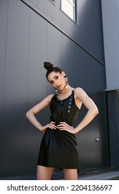 High End Fashion Image Of Tall Slim Brunette Model Wearing Black Leather Dress. Statement Sequins Earrings And Bright Vivid Brown Make Up, Bun Hairstyle. Outdoor Photo Shoot For Summer Campaign