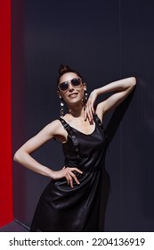 High End Fashion Image Of Tall Slim Brunette Model Wearing Black Leather Dress. Statement Sequins Earrings And Bright Vivid Brown Make Up, Bun Hairstyle. Outdoor Photo Shoot For Sunglasses Campaign