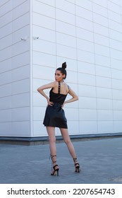 High End Fashion Image Of Tall Long Legs Slim Brunette Model Wearing Short Black Leather Dress, High Heels Summer Shoes. Bun Hairstyle. Outdoor Photo Shoot Over White Wall Background With Copy Space